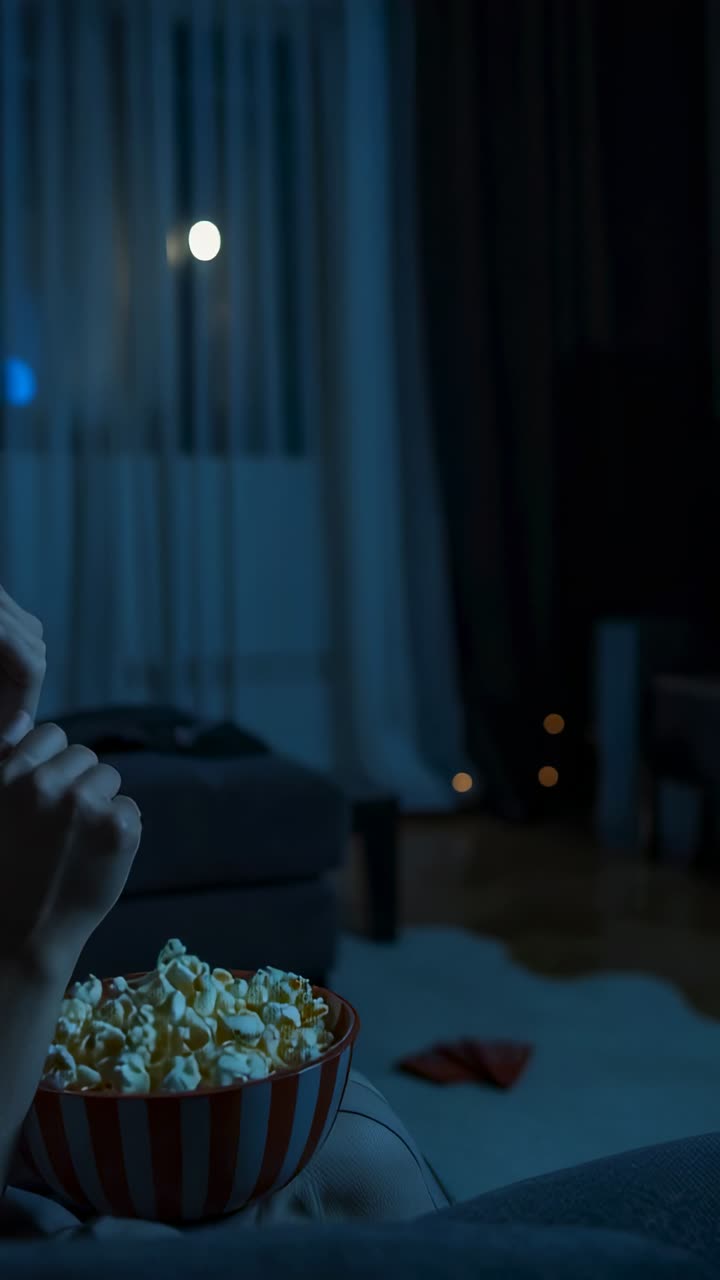 Vertical video: Munching woman raising popcorn from striped bowl on sofa at home with paper plane