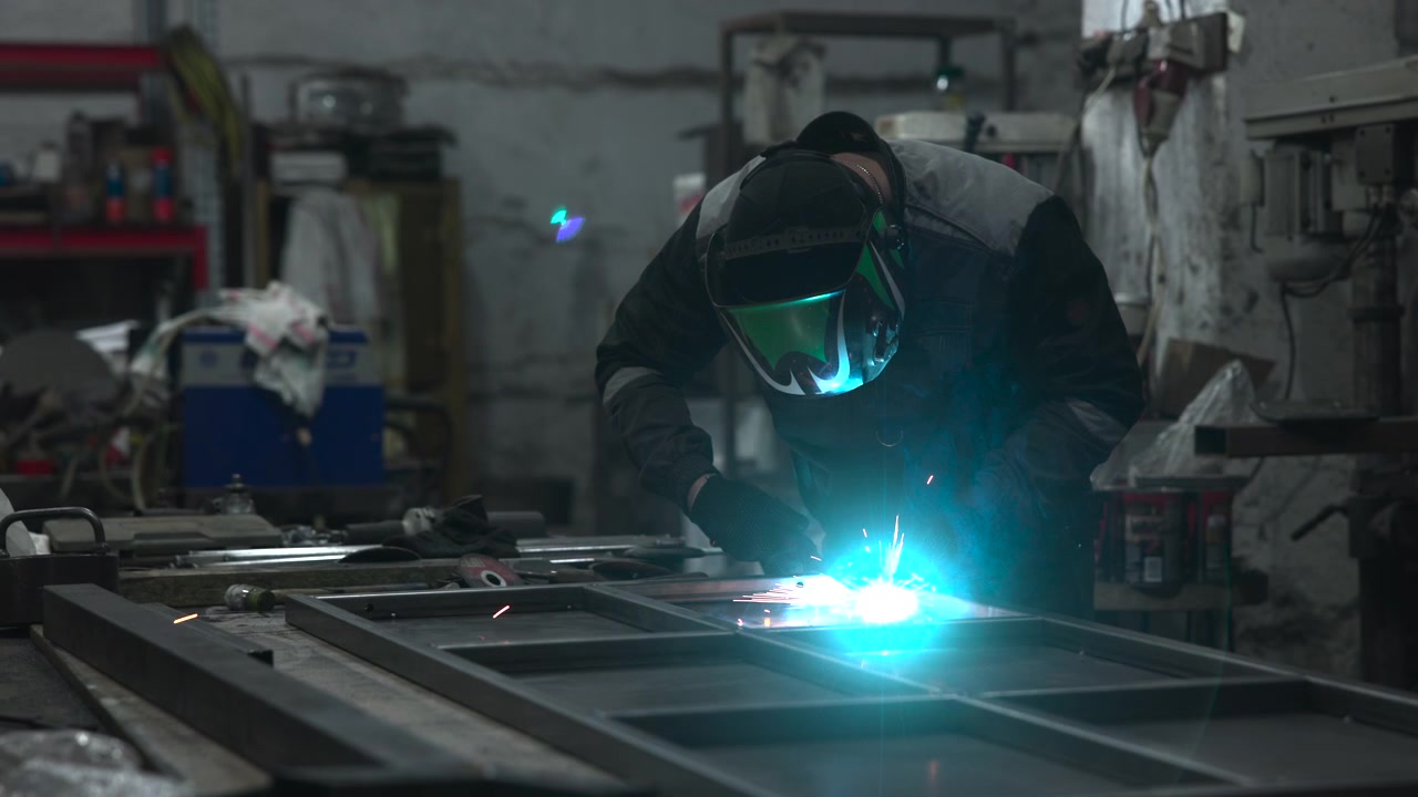 Worker in dark protective jacket and gloves leans over steel sections during welding, creating bright orange sparks, light smoke, and focused workshop atmosphere around fabrication tools