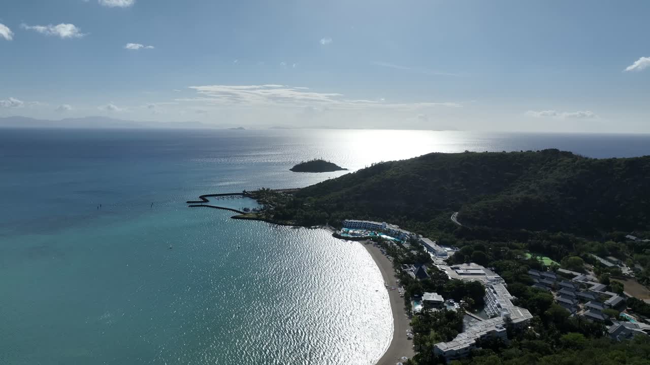 Pomalý odpolední let dronu s jemným panorámováním doleva, který se vznáší nad zálivem Hayman Island. Do záběru se dostává třpytící se sluncem osvětlená voda a pobřežní ostrůvek. Whitsundays, Queensland, Austrálie.