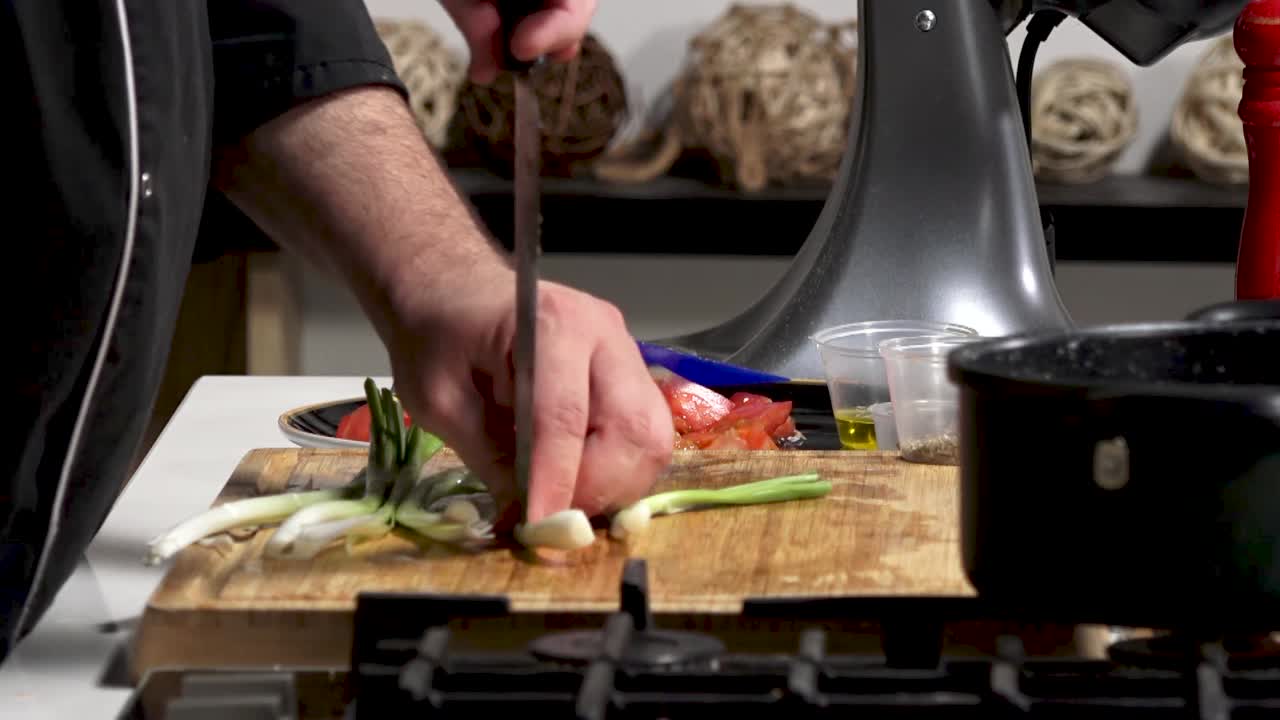 An experienced cook chops fresh vegetables like tomatoes and green onions in a bright kitchen. The chef showcases knife skills while preparing for a tasty dish