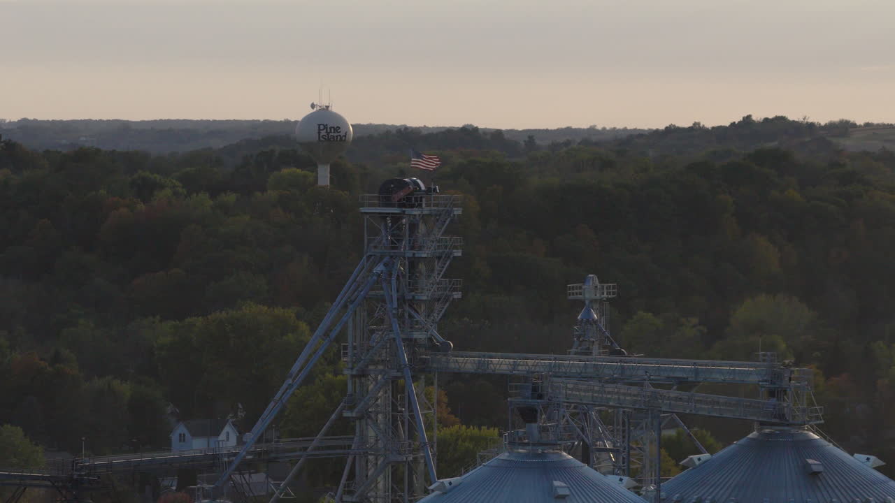 Torre de água e estruturas de elevador de grãos acima das árvores da cidade em Pine Island, Minnesota, com drone aéreo acompanhando para a esquerda enquanto a bandeira dos EUA tremula, destacando a agricultura americana