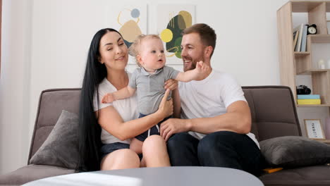 Loving-Parents-Sitting-On-Sofa-In-Living-Room-With-Their-Adorable-Little-Boy-While-Looking-At-Camera-During-A-Video-Call-At-Home-1