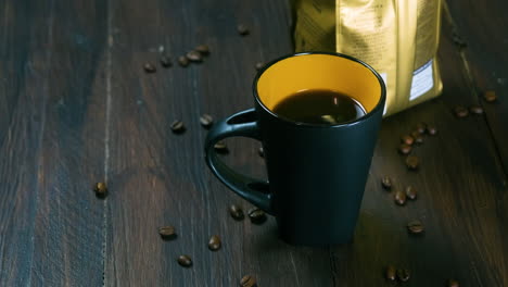 hand of a black man puting down a cup of steaming coffee next to a packet of coffee beans on a wooden table