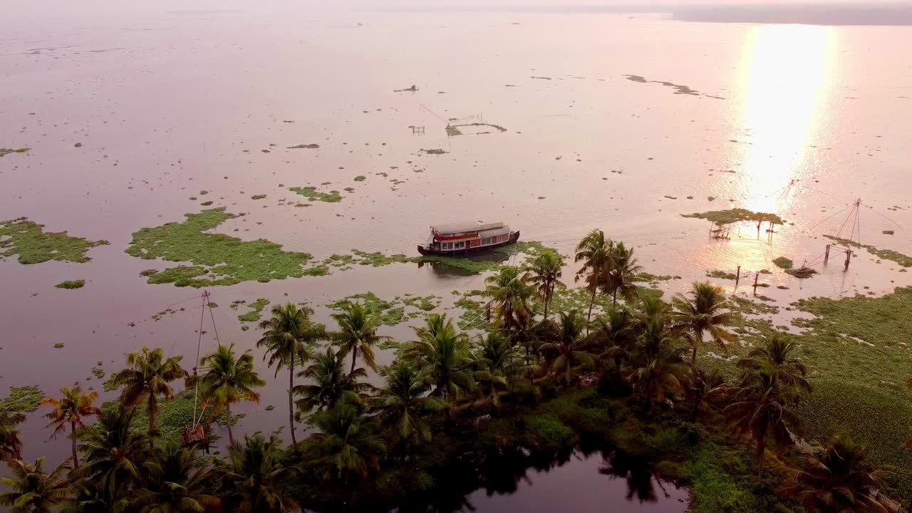 Ripresa aerea con drone di una tradizionale casa galleggiante che naviga al tramonto nelle calme acque del Kerala, circondata da palme da cocco e dalla lussureggiante vegetazione tropicale dell'India meridionale