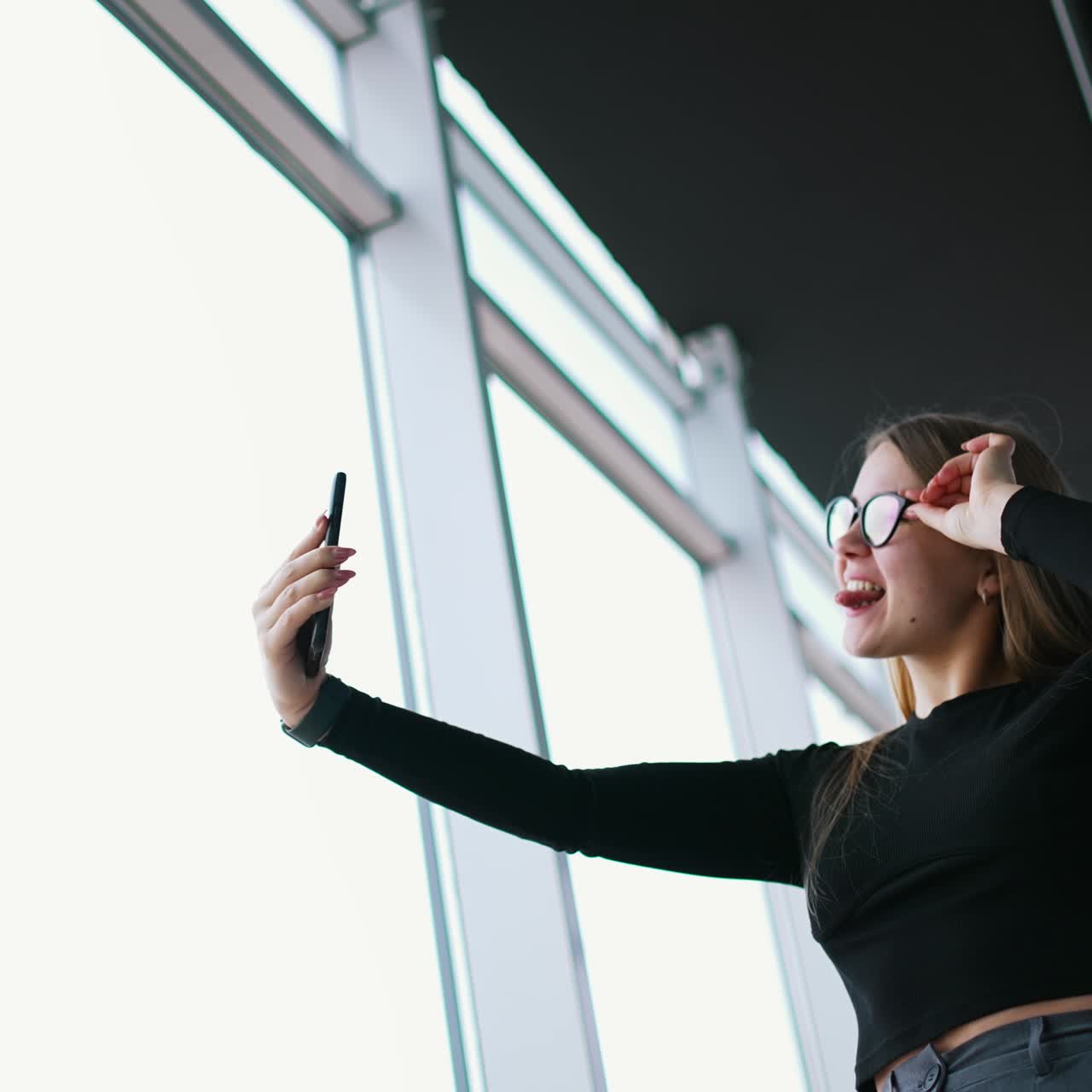 Attractive young woman in stylish glasses with a phone. Beautiful girl holding smart phone doing a selfie photo and smiles showing tongue. Panoramic windows background in office