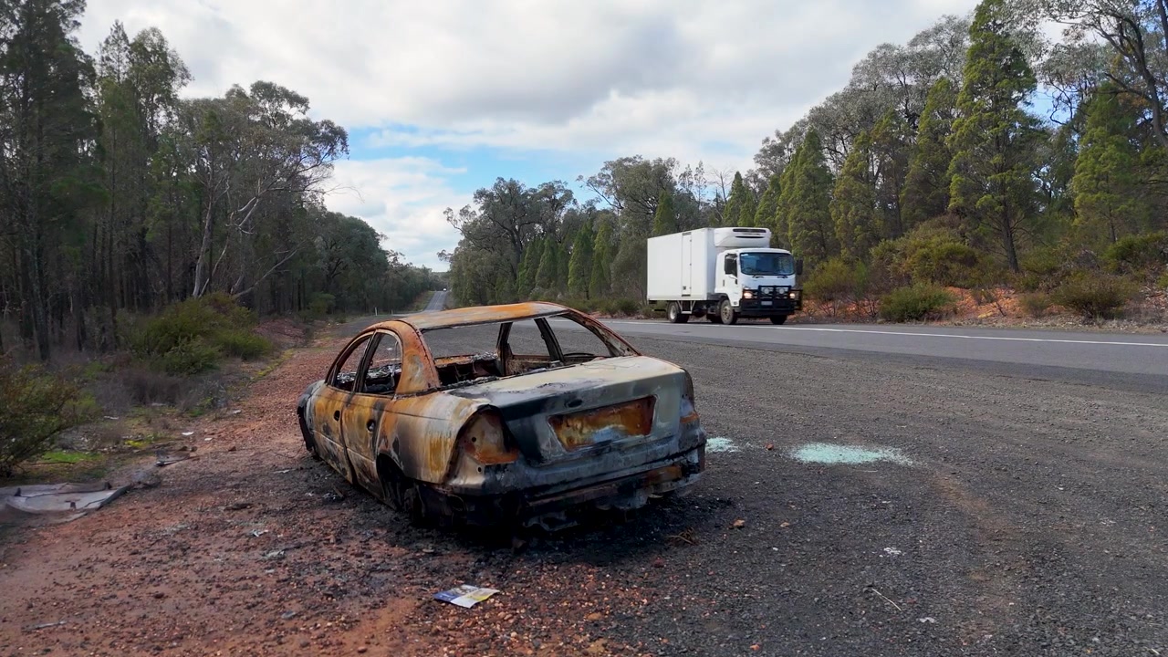 Een verkoold autowrak staat op de vluchtstrook van een snelweg terwijl een vrachtwagen passeert in Australië.