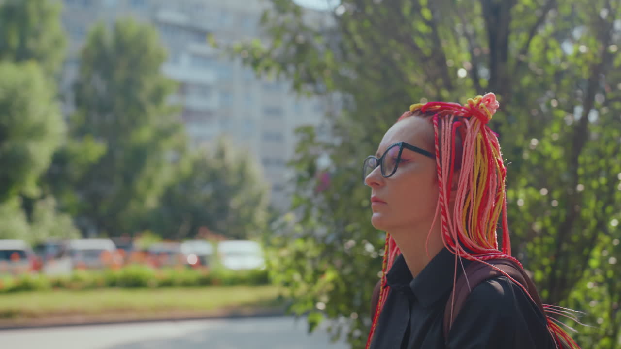 Mujer caucásica pensativa con trenzas rosas de pie en la calle de un apartamento, edificios de la ciudad y árboles al fondo, gafas, mochila, luz suave del día, estado de ánimo reflexivo, retrato urbano solitario que transmite