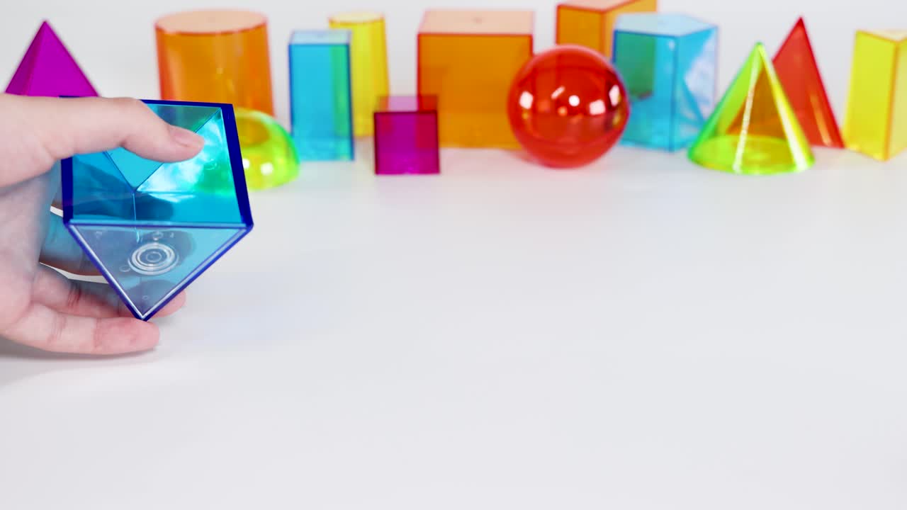 Hands manipulate colorful 3D geometric shapes on a white table, showcasing educational and playful interaction with prisms and pyramids