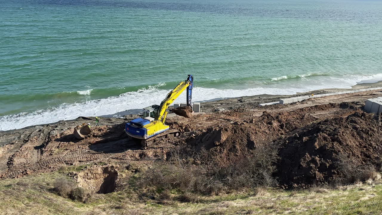 Construction equipment works on coastal land near the water during daylight hours to prepare for a new project close to the shoreline