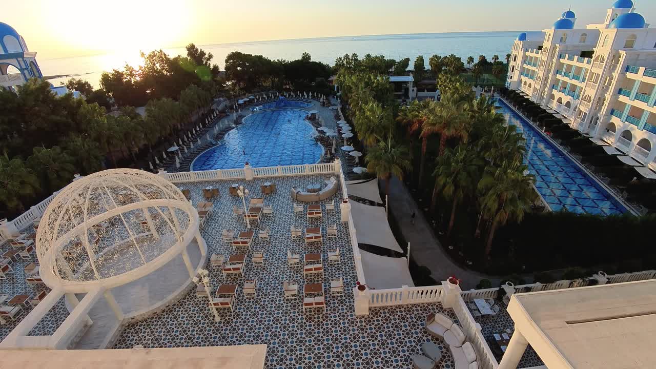 Aerial view of a luxury resort at sunrise with a large pool, palm trees, and white buildings overlooking the ocean, showcasing a beautiful, relaxing holiday destination.