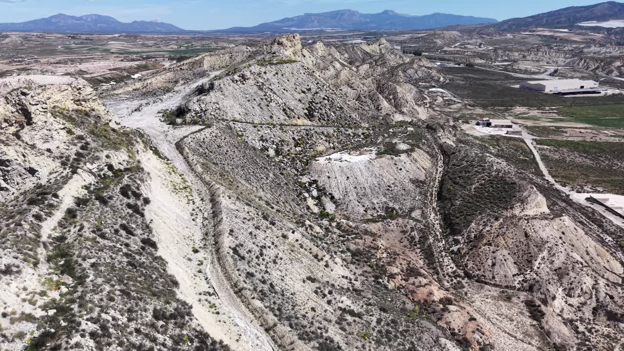 Una ripresa con drone in tracking che si muove attraverso le aride e erose badlands di Lorca, mostrando creste ripide e un deposito d'acqua vicino a siti industriali.