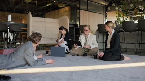 Zoom-in-group-of-confident-businesswomen-in-business-clothes-talking-while-sitting-on-a-gray-sofa-while-a-brunette-businesswoman-girl-breastfeeds-her-small-child