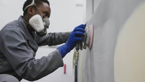 african american male car mechanic wearing a face mask and using a grinder on the side of a car