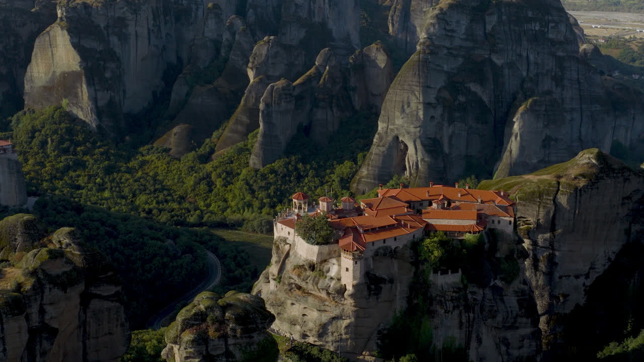 Cinematic aerial view of Meteora monastery in Greece perched on towering cliffs, dramatic rock formations and lush green valley create a breathtaking historic scene
