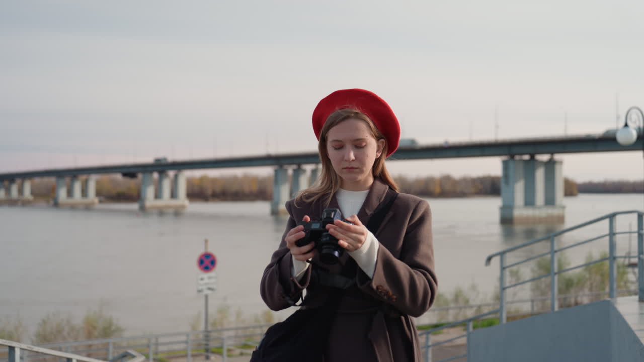 Donna caucasica che controlla le impostazioni della fotocamera, regola l'obiettivo, testa l'esposizione, compone un panorama fluviale, con concentrazione intensa. Dettagli di berretto e cappotto, luce soffusa, sessione pratica di fotografia in città.