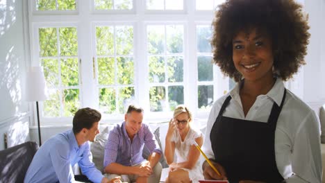 Portrait-of-smiling-waitress-with-notepad-while-friends-interacting-in-background