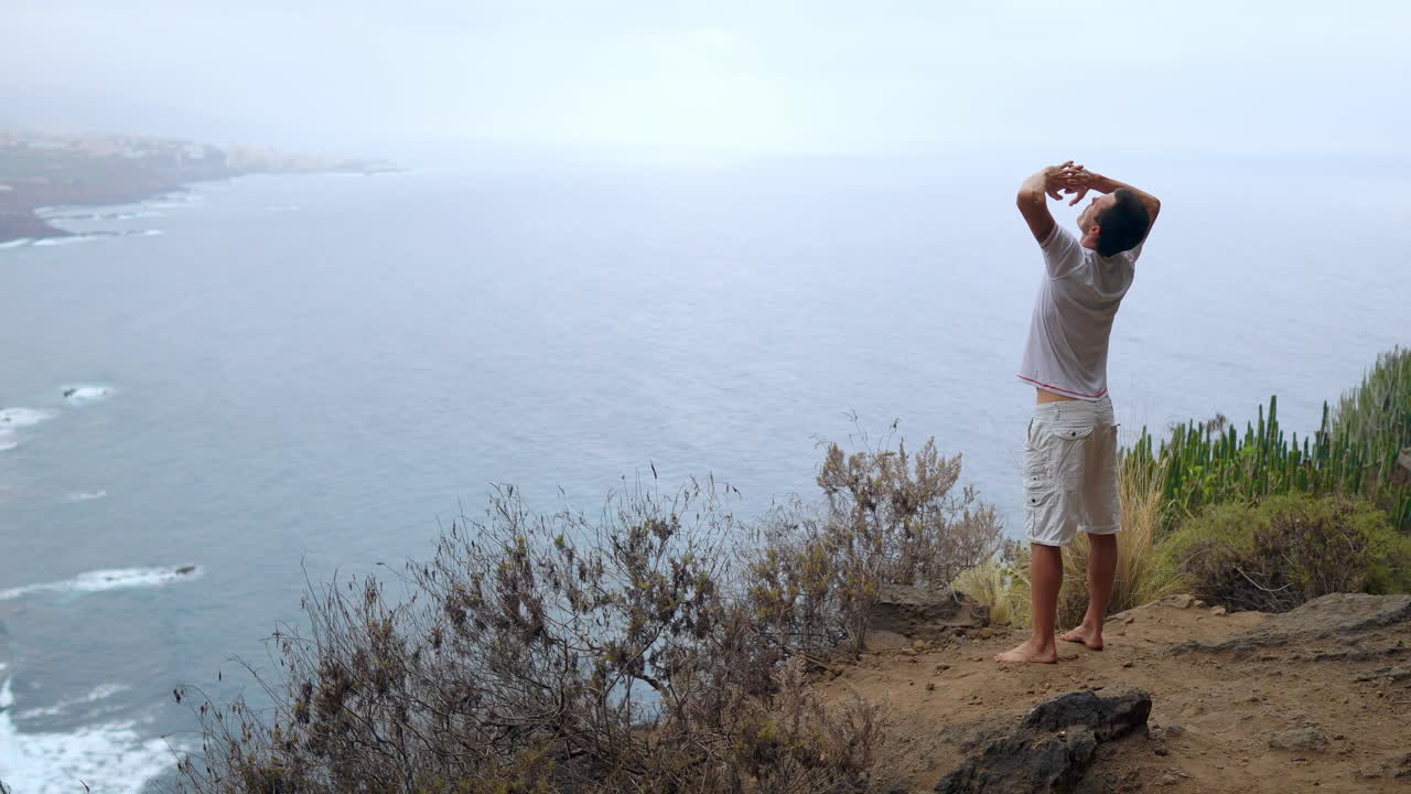 On a cliff's edge, a man extends his arms and breathes in the ocean air during a yoga session, absorbing its tranquility