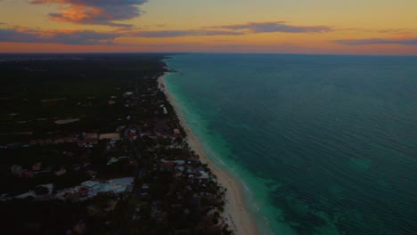 Drone-shot-of-the-green-sea,-with-the-sandy-Akiin-beach-and-the-houses-between-the-trees,-at-sunset