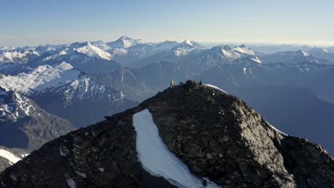 Man-standing-on-the-summit-of-a-huge-mountain-with-rock-and-snow-on-the-peak-amongst-a-spectacular-mountain-range-high-above-the-valleys-in-New-Zealand-during-sunrise