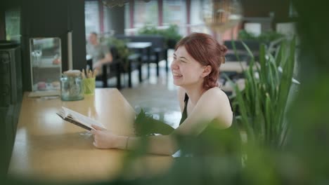 young woman reading a book and sipping a cocktail with a straw at a wooden counter in a bright, modern cafe with natural light. relaxed and focused ambiance