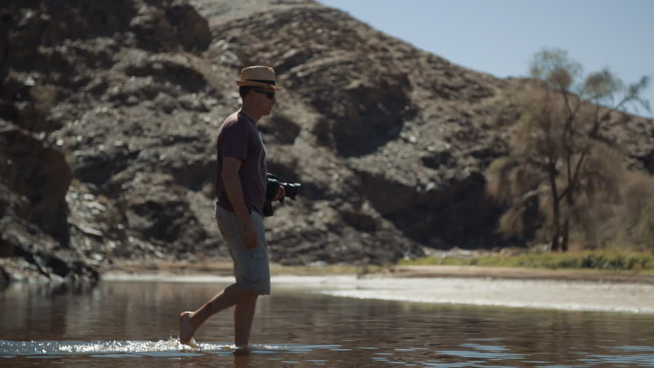 Handheld profile wide slow-motion shot of a photographer tourist with a stylish hat walking through a river as the camera pans with him as he carries a camera.