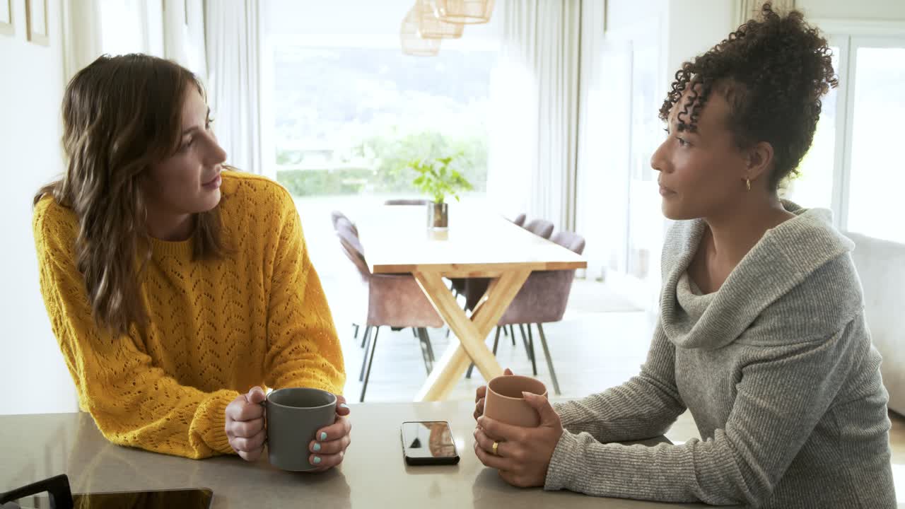 Diverse female friends leaning forward at kitchen counter chatting and catching up over coffee mugs