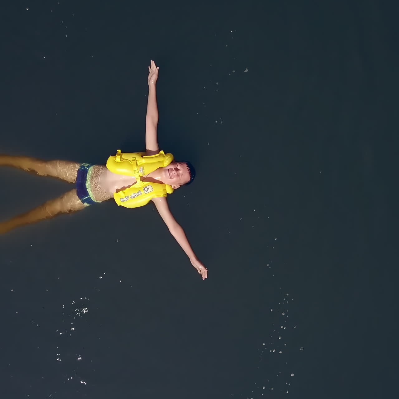 Happy little child in yellow life vest swimming alone on his back in the calm river. Teenage boy wearing safety waistcoat cheerfully swimming. Aerial view. Motion bottom up