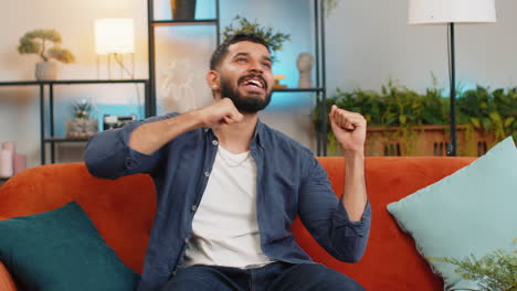 Energetic-happy-Indian-man-singing-dancing-with-hands-listening-music-sitting-on-couch-in-home-room