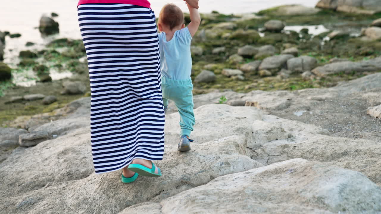 Lovely toddler boy in blue clothes stepping by the grey stones. Mom in long striped skirt supporting her child by the hands. Waterfront at backdrop.