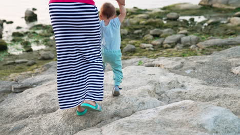 lovely toddler boy in blue clothes stepping by the grey stones. mom in long striped skirt supporting her child by the hands. waterfront at backdrop.
