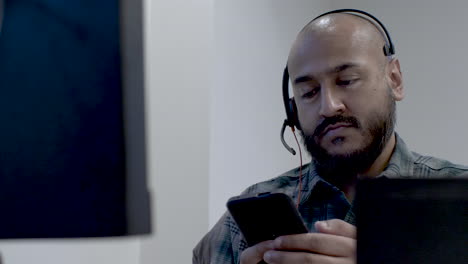 Close-up-shot-of-an-Asian-call-centre-employee-taking-a-break-from-calls-while-sitting-at-his-workstation,-the-man-focused-on-his-mobile-phone-as-he-responds-to-messages-while-swaying-back-and-forth