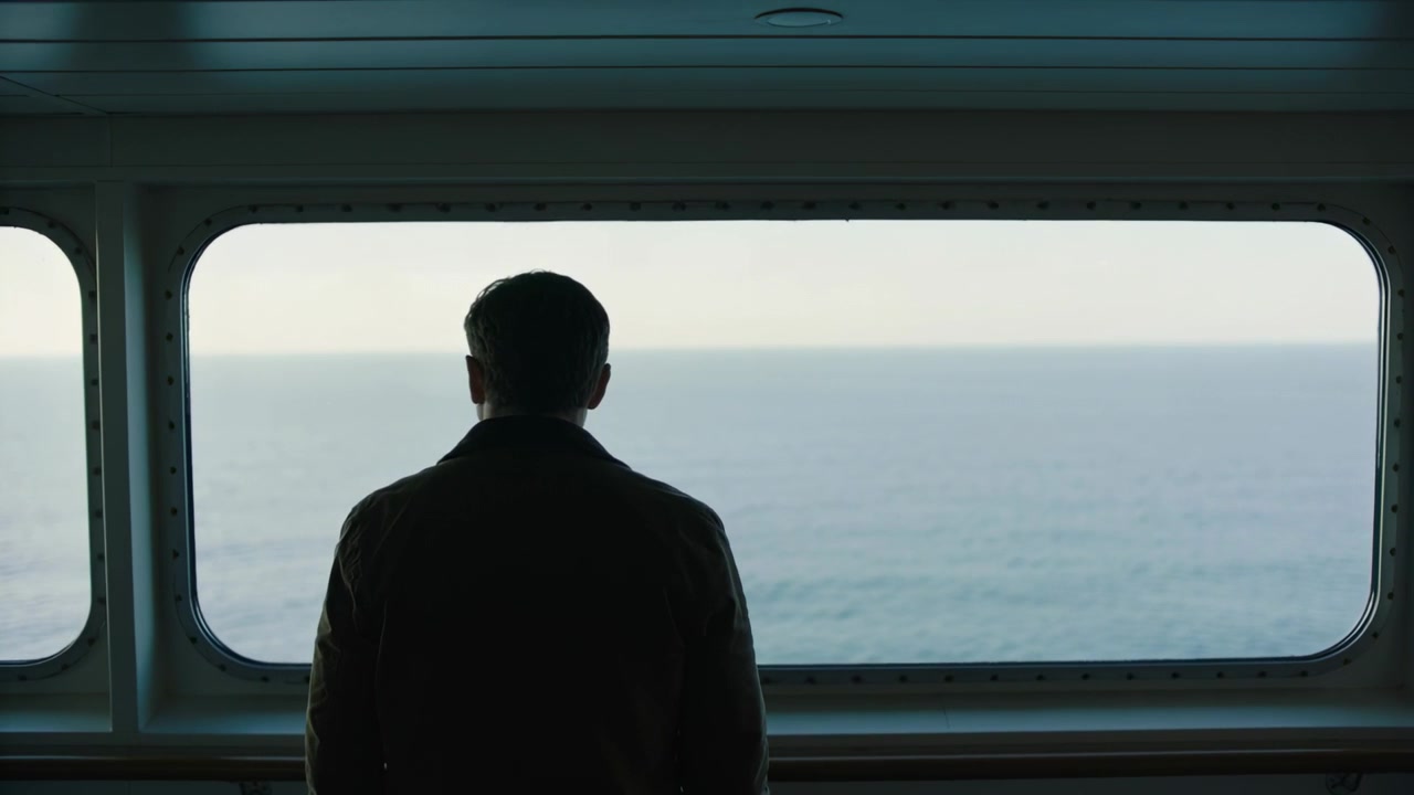 Ship leaving port causing male passenger standing at large ship window ledge, gazing at calm sea