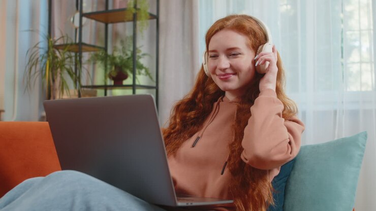 Felice giovane donna caucasica rilassata con le cuffie wireless che ascolta musica usando il portatile sul divano