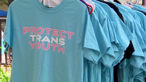 Un-Primer-Plano-Medio-De-Un-Estante-De-Camisetas-Azules-Con-El-Logotipo-De-Protección-De-La-Juventud-Trans-Con-Diversidad,-Equidad-E-Inclusión