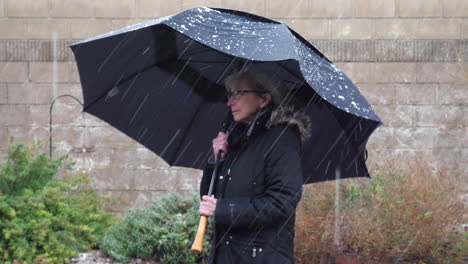 Una-Anciana-Con-Un-Paraguas-Negro-Y-Un-Abrigo-De-Lluvia-En-Un-Mal-Tiempo-Helada-Tormenta-De-Invierno-Mientras-La-Nieve-Y-El-Granizo-Caen-Alrededor-De-Su-Cámara-Lenta