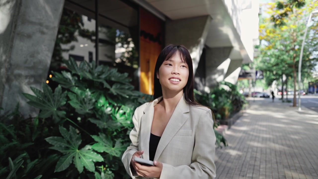 微笑的亚洲年轻女性手持手机伫立城市人行道