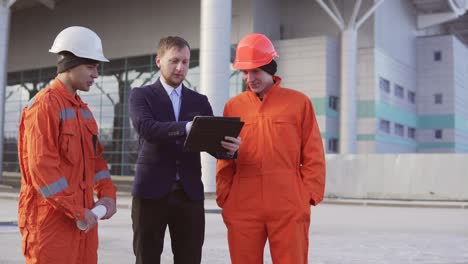 Investor-of-the-project-in-a-black-suit-examining-the-building-object-with-construction-workers-in-orange-uniform-and-helmets.-They-are-cheking-the-drawings-using-the-tablet.