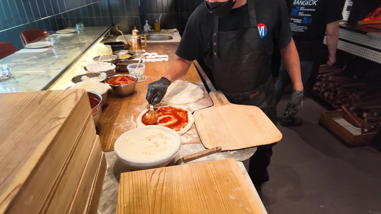 Chef professionista con grembiule nero spalma salsa di pomodoro su impasto di pizza crudo sul bancone della cucina.