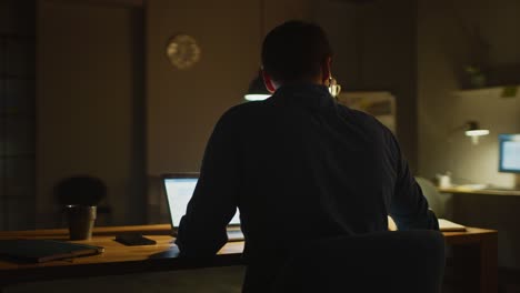 professional creative man sitting at his desk in office studio working on a laptop in the evening. man working with data and analyzing statistics, using smartphone. 360 degree tracking arc shot movement