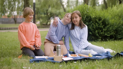 Kleines-Mädchen-Mit-Down-Syndrom-Und-Ihre-Freunde-Lächeln-In-Die-Kamera,-Während-Sie-Im-Park-Auf-Dem-Gras-Sitzen