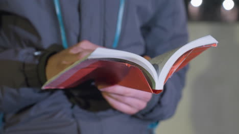 close-up hand view of man flipping red cover book outdoors at night with blurred bokeh effect from passing car lights, illuminated by urban city lights