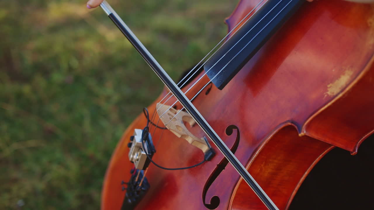 Female hand holding a bow and playing cello instrument outdoors. Close up. Green grass backdrop in blur.