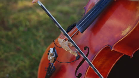 female hand holding a bow and playing cello instrument outdoors. close up. green grass backdrop in blur.