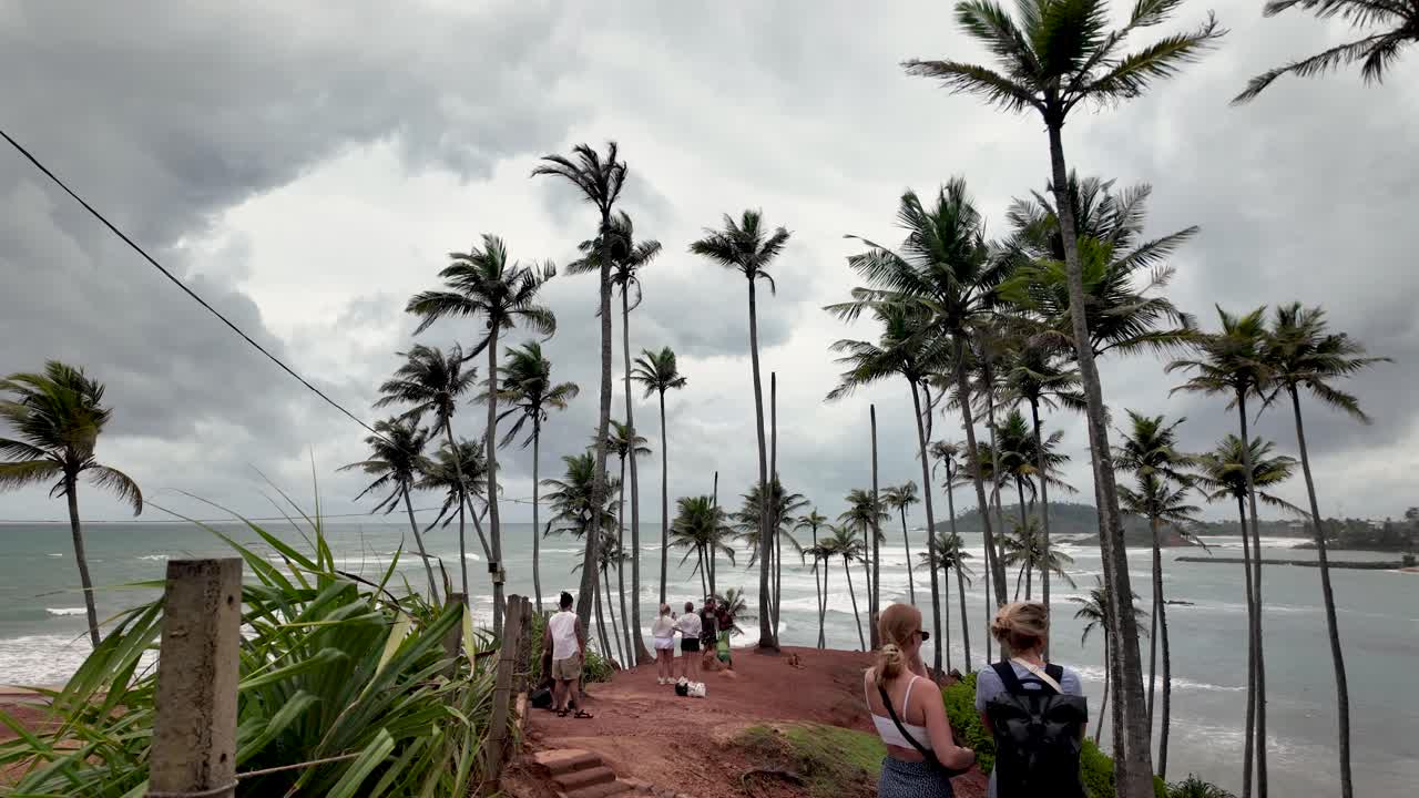 Tourists admire the view at Coconut Tree Hill in Mirissa, Sri Lanka, surrounded by tall palm trees and overlooking the ocean on a cloudy day.