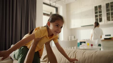 happy man playing with his daughter who is cheerfully balancing on his arms in airplane pose while mom is busy in the kitchen, on a light brown sofa in a studio apartment