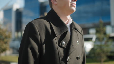 close-up of elegant black coat with shiny metallic buttons, paired with briefcase held firmly, in outdoor urban setting featuring blurred cars, greenery, and glass buildings