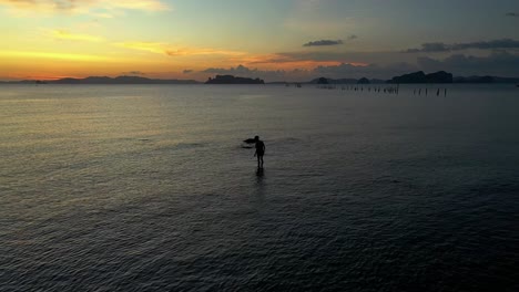 the silhouette of a young boy wandering and stumbling in the shallow sea waters of scenic coastline during sunset, aerial pullback shot