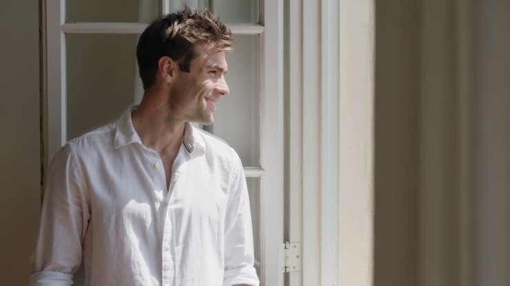 Notando la luce del sole, un uomo in camicia bianca si gira e sorride in una stanza luminosa, guardando fuori dalla finestra aperta.