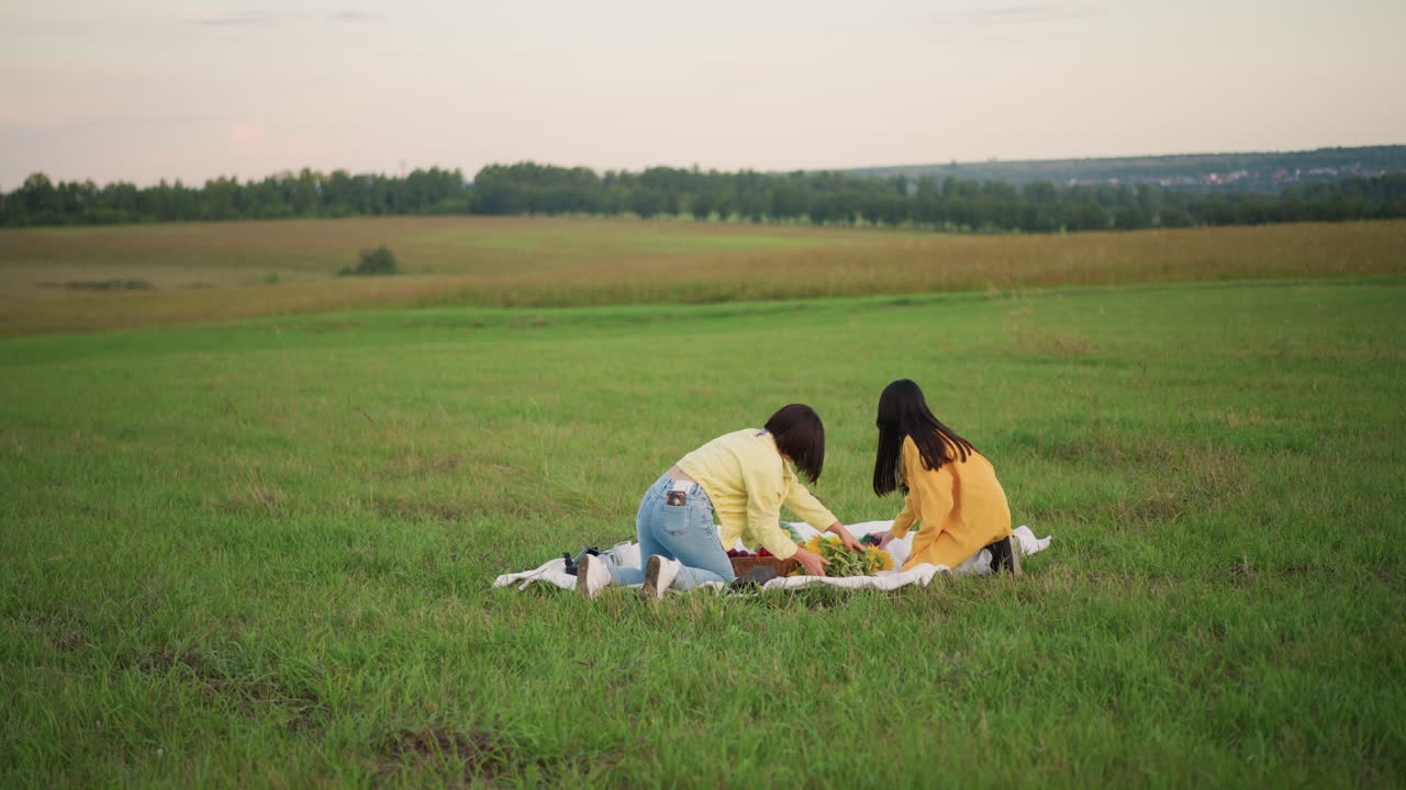 해 질 녘, 초원에서 피크닉을 즐기는 두 명의 아시아 여성. 대학생들이 담요 위에서 스케치를 하고 간식을 나눠 먹고 있어요. 데님 재킷과 노란색 재킷, 부드러운 빛, 넓은 시골 지평선, 차분한 대화, 편안한 분위기, 친밀함.