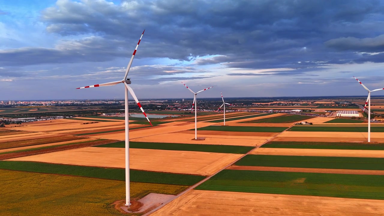 Vindmøllepark blandt geometriske landbrugsmarker. Luftfoto viser hvide vindmøller, markveje og en fjern by under lagdelte skyer